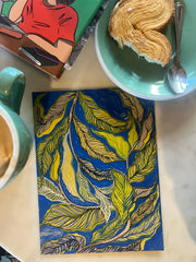 Overhead cafe scene with an illustrated book and pastry, beside a painting of yellow-green leaves on royal blue