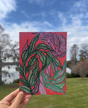 Hand holding a painting of green, pink and purple feather-like leaves on red, outdoors by a lake