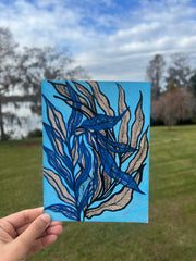 Hand holding a painting of navy blue and cream feather-like leaves on turquoise, outdoors with trees and sky